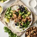 A bowl filled with grilled chili-lime chicken, white rice, black beans, avocado salsa, and fresh cilantro, garnished with lime wedges and green onions.