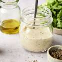 Jar of creamy Greek salad dressing with herbs, olive oil, and fresh lettuce in the background.