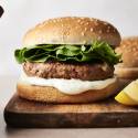 Greek turkey burger on a wooden board with crisp lettuce and creamy tzatziki sauce inside a sesame seed bun, garnished with a lemon wedge on the side.