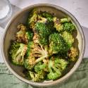 A bowl of roasted broccoli with crispy edges, sprinkled with red pepper flakes, sits on a light surface beside a salt dish.