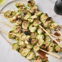 Grilled Brussels sprouts skewered and arranged on a white platter, drizzled with balsamic glaze and sprinkled with cracked black pepper, with rosemary and olive oil in the background.