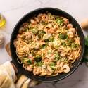 A skillet of shrimp and broccoli linguine, garnished with parsley and Parmesan. Surrounding are garlic cloves, olive oil, and a pepper mill on a marble surface.