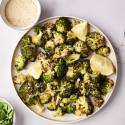 A plate of roasted broccoli garnished with lemon wedges, surrounded by parsley, grated cheese, and a pepper grinder.
