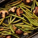 Roasted green beans and mushrooms on a baking sheet with sliced garlic and balsamic vinegar.