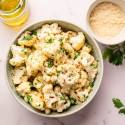 A bowl of roasted cauliflower florets garnished with chopped parsley on a marble surface. Nearby are a bowl of grated cheese and a jar of olive oil.