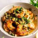 A bowl of golden-brown shrimp coated in a lemony garlic butter sauce, garnished with capers and fresh herbs. The setting conveys a fresh, appetizing tone.