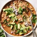 Slow cooker chicken enchilada soup in a white bowl, garnished with avocado, jalapeños, red onion, cilantro, green onions, and shredded cheese, served with a spoon.