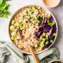 A colorful bowl of brown rice topped with cubed avocado, diced cucumber, purple cabbage, and dressing. Celery and a linen cloth are nearby, creating a fresh and vibrant look.