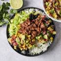 A vegan ground beef burrito bowl is served with cilantro lime rice, black beans, lettuce, seasoned plant-based ground beef, avocado, corn, and fresh pico de gallo, garnished with chopped cilantro and a lime wedge.