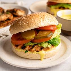 A grilled chicken burger with lettuce, tomato, pickles, and mustard in a sesame bun on a plate. Grilled chicken and sauce in the background.