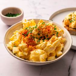 Bowl of egg salad topped with paprika and chopped chives, next to a slice of bread with egg salad. Small bowl of chives in the background.
