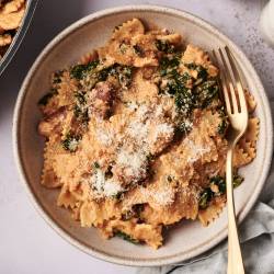 Creamy high-protein pasta made with bowtie noodles, sausage, spinach, and ricotta, served in a beige bowl and topped with grated Parmesan cheese. A gold fork rests on the side.  