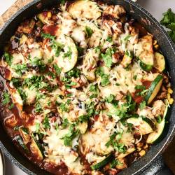 A skillet of cheesy chicken, black beans, and zucchini is served on plates, garnished with fresh cilantro and spices.