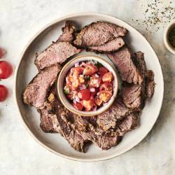 Platter of sliced beef with a bowl of fresh tomato salsa, surrounded by cherry tomatoes and spices on a light background.