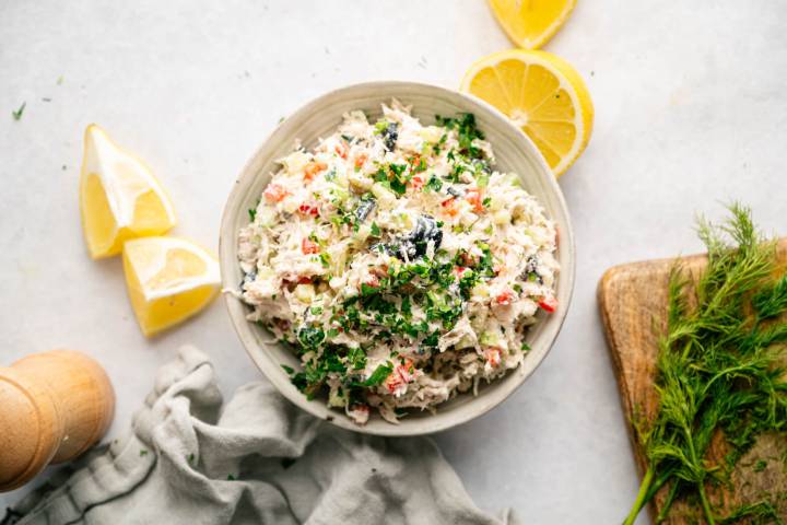 A bowl of colorful crab salad garnished with fresh herbs, surrounded by lemon wedges, dill sprigs, a wooden board, and a gray cloth on a light surface.