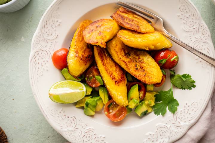 A white plate with golden-brown fried plantains, sliced avocado, cherry tomatoes, lime wedge, and cilantro. The setting is light, fresh, and inviting.