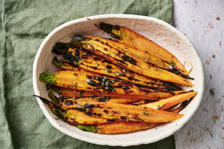 A white bowl filled with charred orange carrots topped with herbs, resting on a green cloth and surrounded by a textured surface.