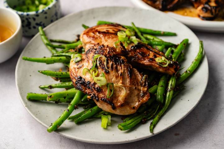 Grilled chicken breast topped with green onions on a bed of green beans on a white plate. A small bowl of sauce sits nearby.