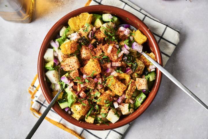 A colorful salad in a brown bowl, featuring diced fruits, veggies, and spices, served with metal spoons on a checkered towel.
