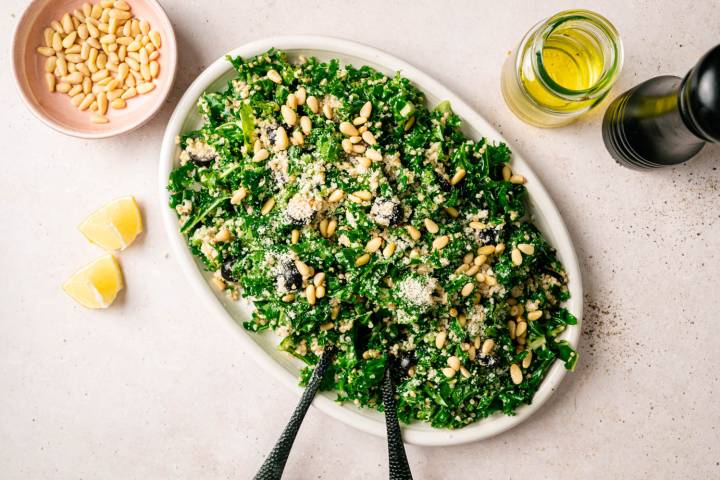 A vibrant kale salad on a white oval plate, garnished with pine nuts, grated cheese, and olives. Nearby are lemon wedges, olive oil, and a pepper grinder.