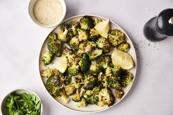 A plate of roasted broccoli garnished with lemon wedges, surrounded by parsley, grated cheese, and a pepper grinder.