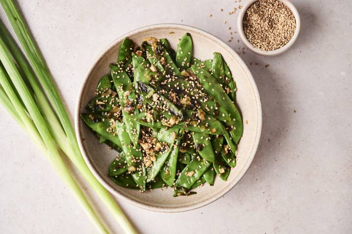 A finished dish of sauteed snow peas topped with sesame seeds in a beige bowl.
