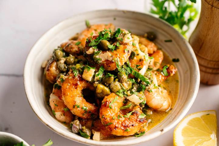 A bowl of golden-brown shrimp coated in a lemony garlic butter sauce, garnished with capers and fresh herbs. The setting conveys a fresh, appetizing tone.