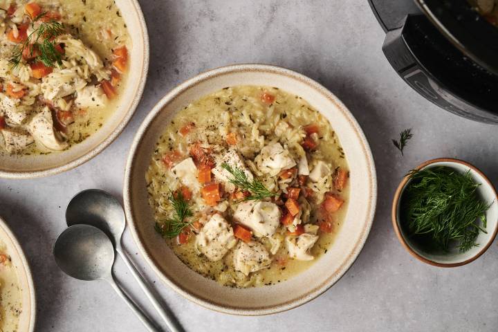 Two bowls of creamy chicken and rice soup garnished with fresh dill and diced tomatoes, accompanied by two spoons and a bowl of herbs.