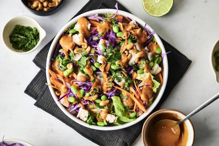 A vibrant salad with purple cabbage, carrots, edamame, chicken, and cilantro, drizzled with peanut sauce and topped with cashews.