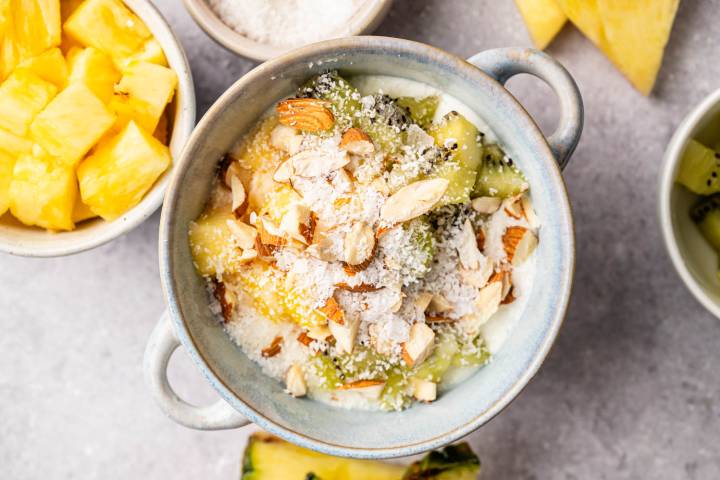 A bowl of yogurt topped with chopped almonds, shredded coconut, kiwi slices, and pineapple chunks. Surrounding it are bowls of pineapple and kiwi on a gray surface.