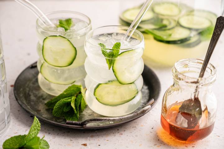 Two glasses of refreshing cucumber mint water with glass straws sit on a metal tray. A jar of honey and a bundle of fresh mint are nearby